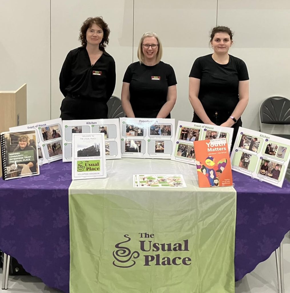 three women dressed in The Usual Place uniform face camera, they are smiling and stand behind an exhibition table for The Usual Place
