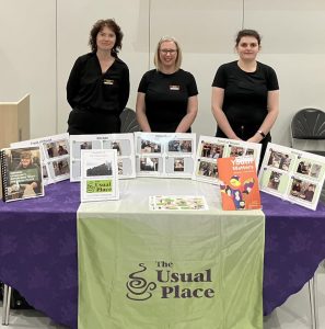 three women dressed in The Usual Place uniform face camera, they are smiling and stand behind an exhibition table for The Usual Place