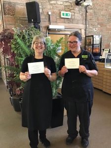 Two Usual Place Mentors stand smiling at the camera and holding up invitations to the Royal Garden Party