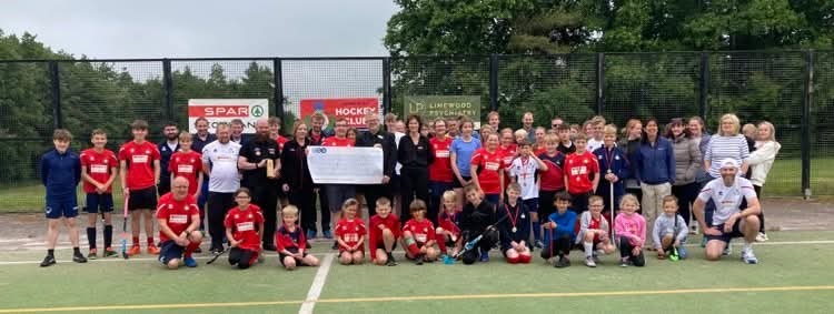 Members of Dumfries Hockey Club of all ages smile at the camera. In the centre is a giant cheque held by The Usual Place CEO Craig and Lived Experience Mentor Kat