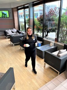 Young woman walks through lounge area of hotel smiling and carrying cups
