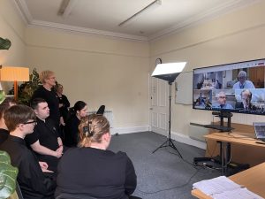 Young people to the left of the image are watching a large screen with members of House of Lords screened on video call