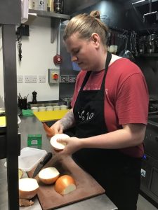 Young man in red tshirt and black 1610 apron peels onions in kitchen