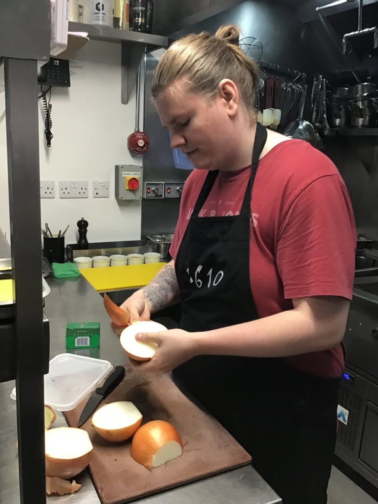 Young man in red tshirt and black 1610 apron peels onions in kitchen