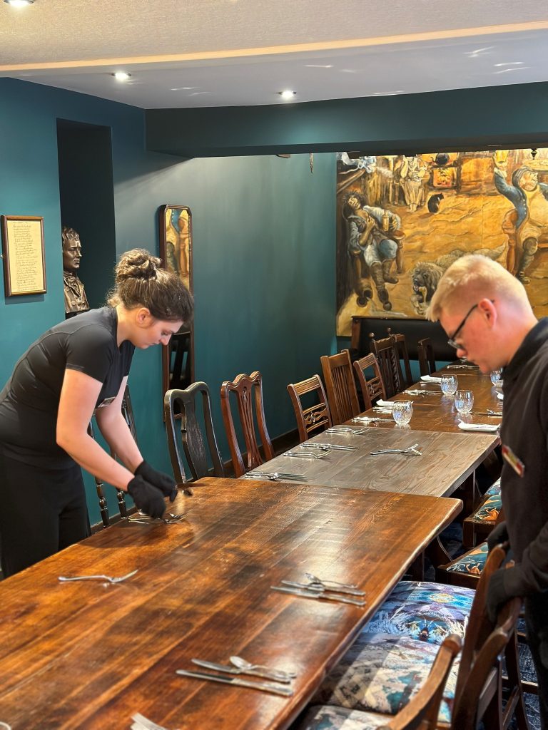 A young woman and young man wearing black serving gloves stand on either side of a table concentrating hard and setting out the cutlery.