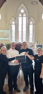 Young man in white shirt stands with three team members from the usual place wearing black. They are all holding a picture of The Cairndale Hotel and Spa logo and smiling