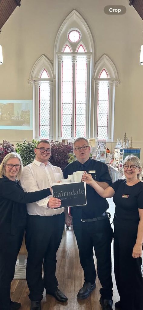 Young man in white shirt stands with three team members from the usual place wearing black. They are all holding a picture of The Cairndale Hotel and Spa logo and smiling