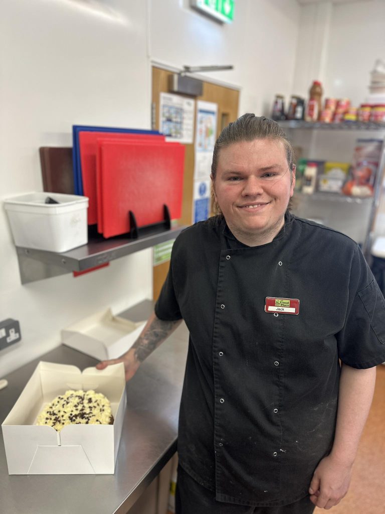 Young chef in kitchen smiling at camera beside him is a highly decorated cake in a box