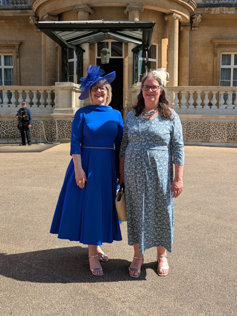 Two ladies dressed in finery, blue tea dresses and stand outside Buckingham Palace in sunshine