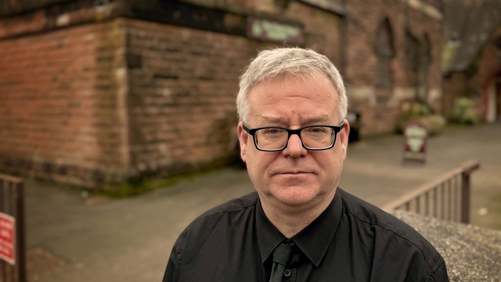 Man with grey hair and glasses dressed in Usual Place blacks looks at camera. In the background is the red sandstone facade of The Usual Place building