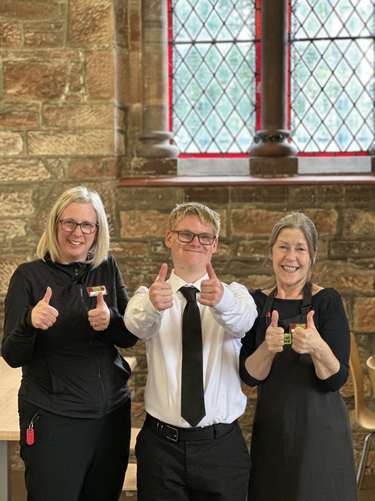 Two women dressed in black unform stand either side of a young man in white shirt and black tie. They are all smiling and doing a thumbs up sign. Behind them is a sandstone wall and ornate window of The Usual Place cafe.