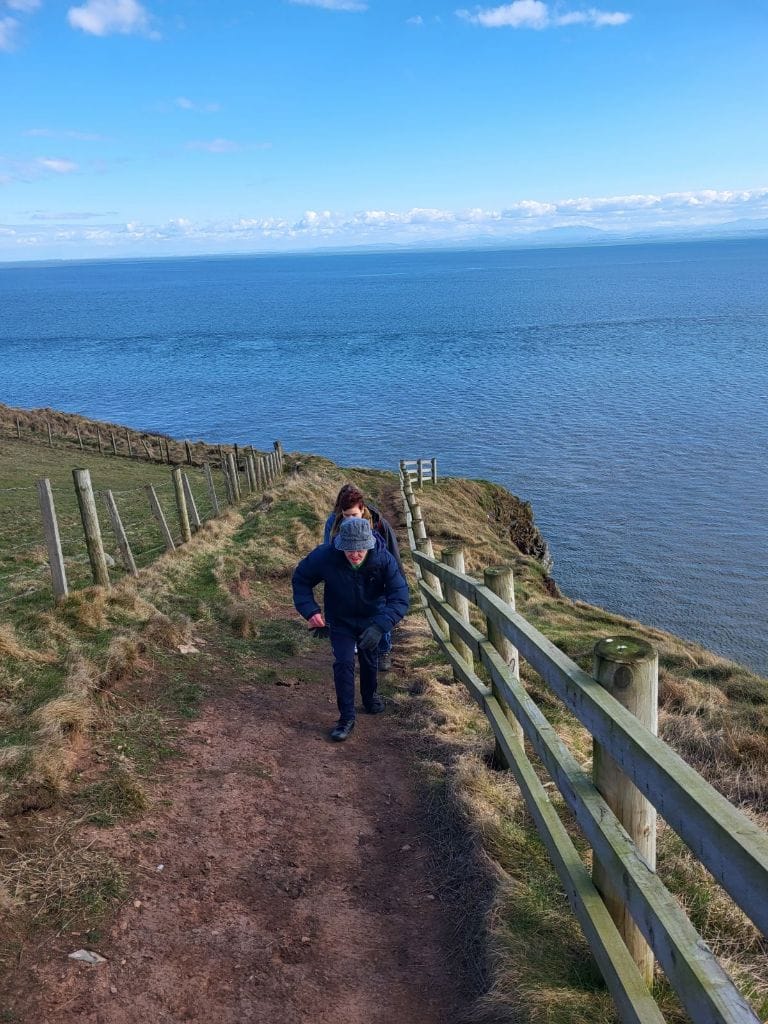 People walking up a steep cliff top with sea behind them and fences on either side