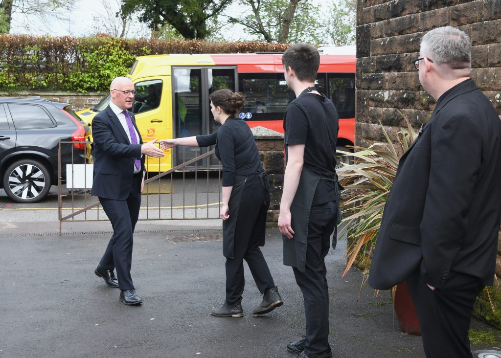 First Minister of Scotland walks towards The Usual Place and is greeted by young woman with outstretched hand. Behind her a young man in Usual Place uniform is waiting to greet him too