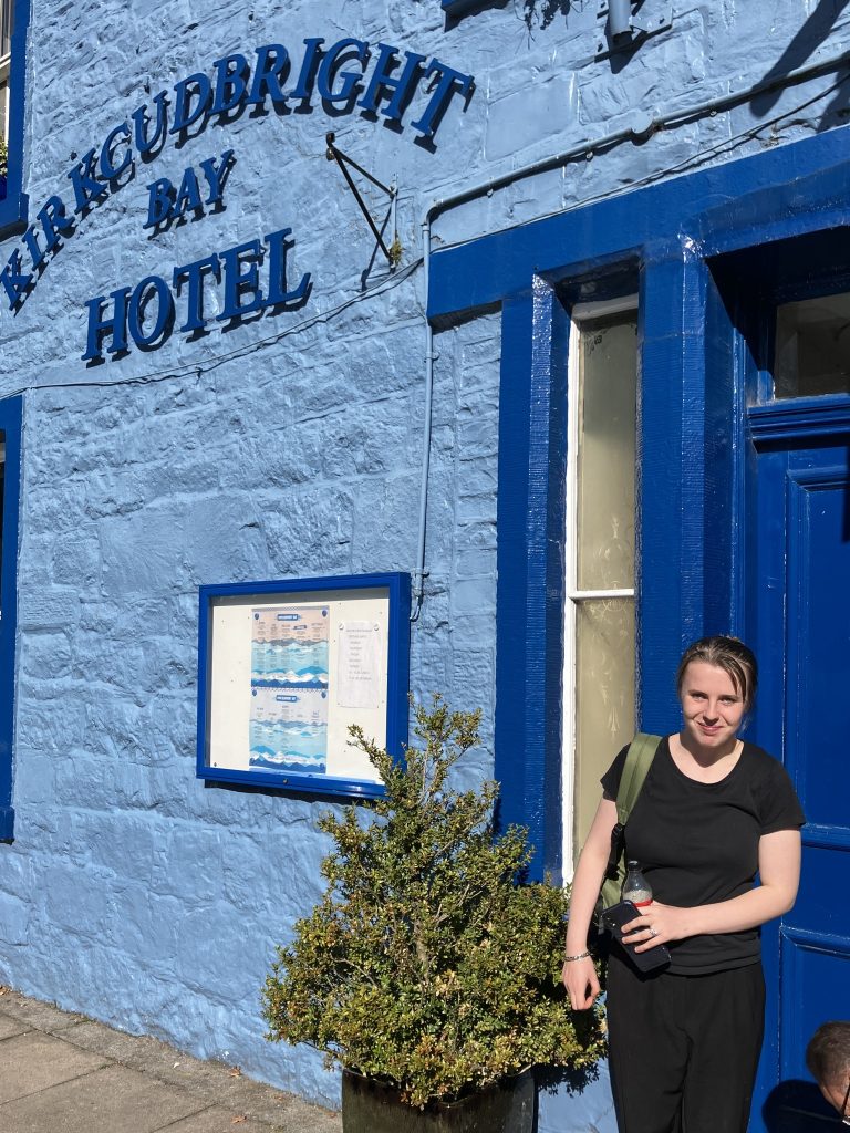 Young woman stands outside the large blue door near blue signage for Kirkcudbright Bay Hotel. She is smiling and holding a bag over her shoulder.