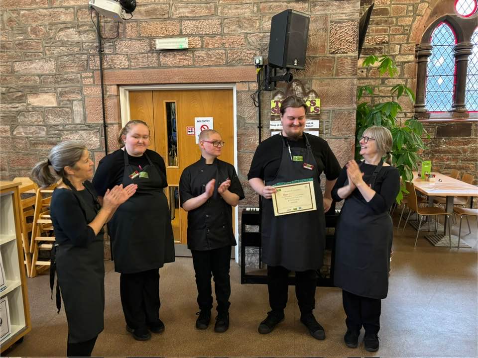 Young man stands and smiles, he is holding a certificate and four people stand around him clapping and smiling