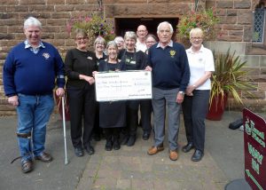 A group of people stand holding a large cheque for £2,000. Some of the people are dressed in Th eUsual Place blacks and the others have Dumfries Lions Club badges on their clothes. They are looking at the camera and smiling outside the entrance to The Usual Place.