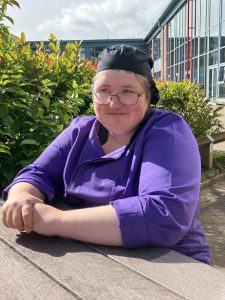 Young woman dressed in chefs hat and purple chef jacket sits in the sunshine at a picnic table. Behind her is the facade of Moffat Academy