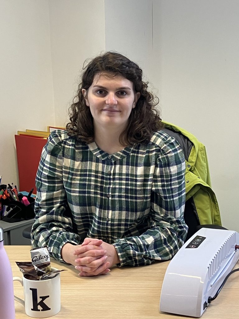 Young woman in checked shirt sits behind a desk and smiles at the camera