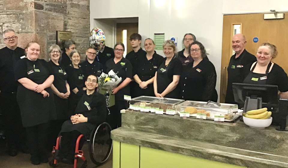 Lady holds flowers and a balloon. She is surrounded by lots of smiling people who are all dressed in The Usual Place Blacks. They are standing to the side of the cake counter with a number of sweet treats displayed