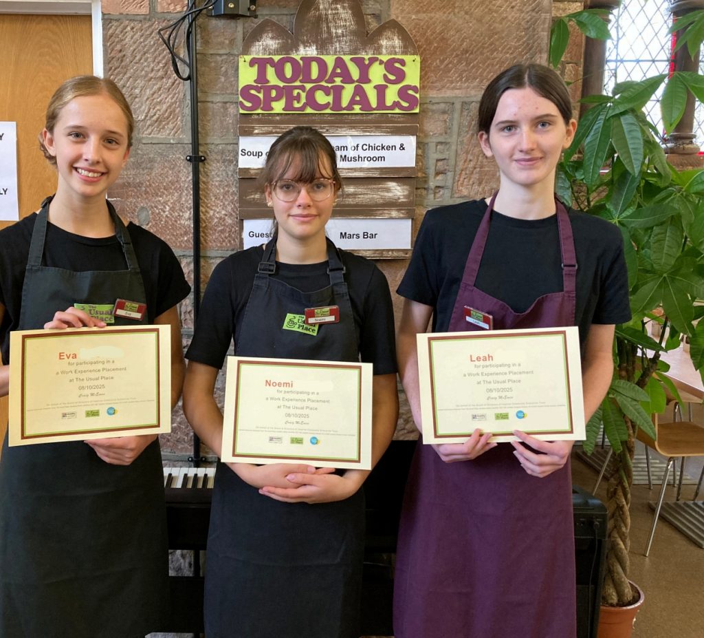3 young girls holding certificates, wearing Usual Place outfits and aprons with the The Usual Place logo. They are looking at the camera and smiling.