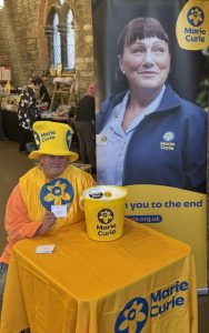 Lady in huge yellow top-hat with blue writing and branded apron and bucket sits behind a table with a large pop up banner for Marie Curie. In the background are people setting up craft stalls.