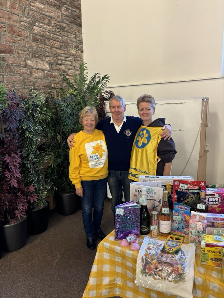 Three people dressed i nbright yellow and blue clothing stand together smiling at the camera, in front of them is part of a tombola stall with lots of ticketed gifts.