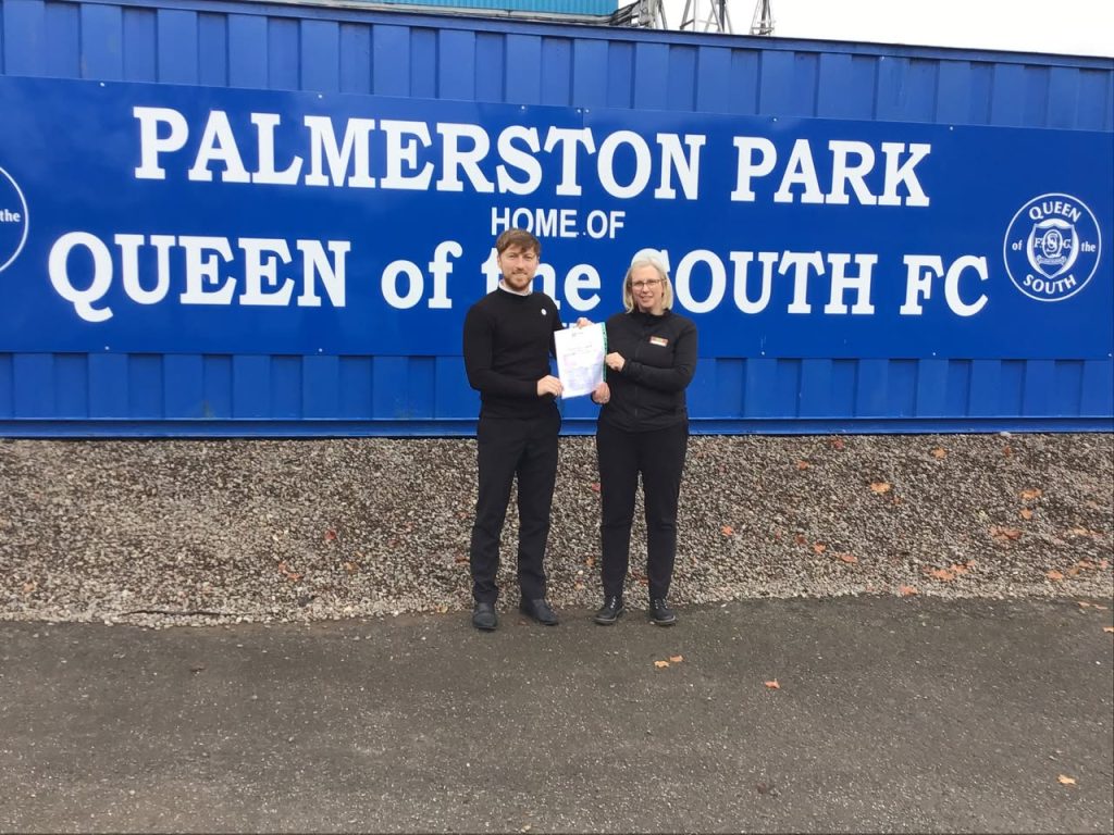 Two people holding a certificate and smiling. They are standing in front of blue signage with white lettering reading Palmerston Park Queen of the South FC