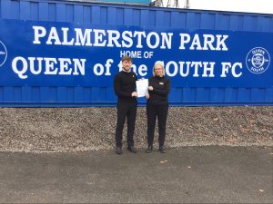 Two people holding a certificate and smiling. They are standing in front of blue signage with white lettering reading Palmerston Park Queen of the South FC