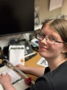 Young woman smiles up at the camera over her shoulder. She is sitting at a desk and using a paper-cutter to finish off some menus she has created at her desk