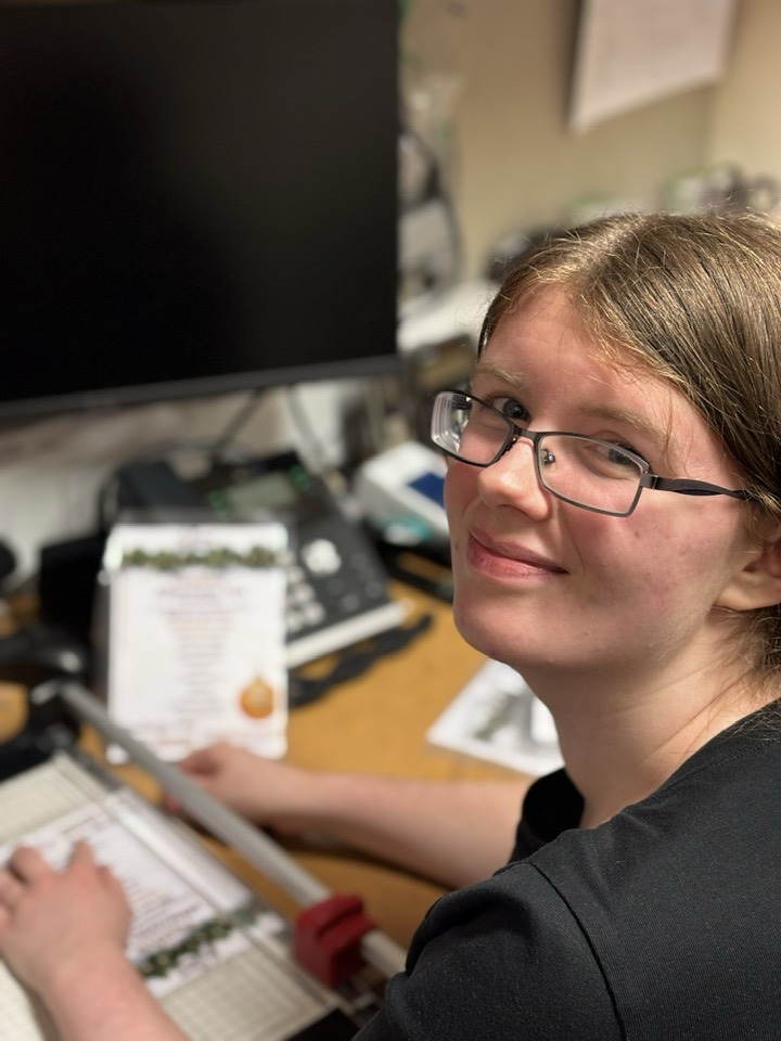 Young woman smiles up at the camera over her shoulder. She is sitting at a desk and using a paper-cutter to finish off some menus she has created at her desk