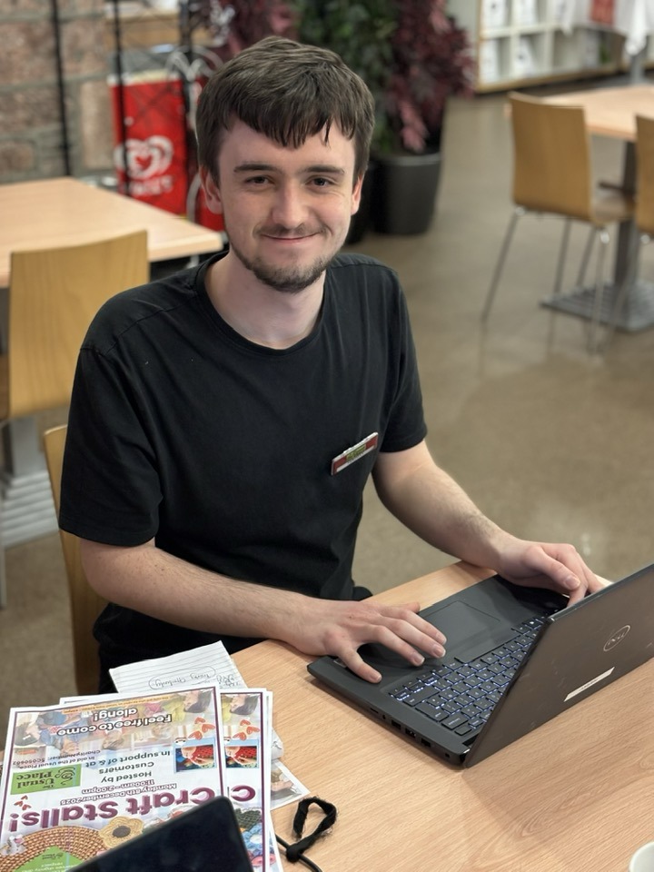 Young man looks at the camera and smiles. He is sitting at a laptop in the cafe at a table. There are drafts for posters beside him - he is making these using his laptop