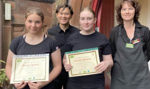 4 people standing and smiling. 2 volunteers in the centre-left foreground holding certificates and 2 mentors in the centre-right background behind them.