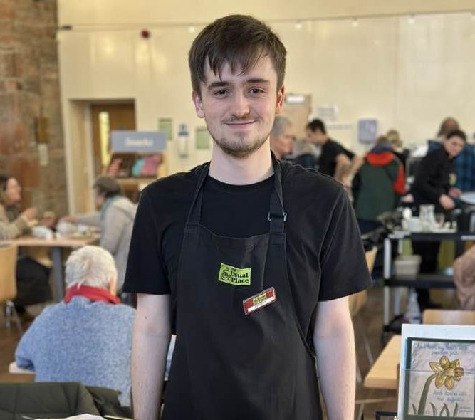 A volunteer standing centre of the photo wearing The Usual Place uniform. He is smiling and standing behind the Welcome board, ready to greet customers and show them to their tables.