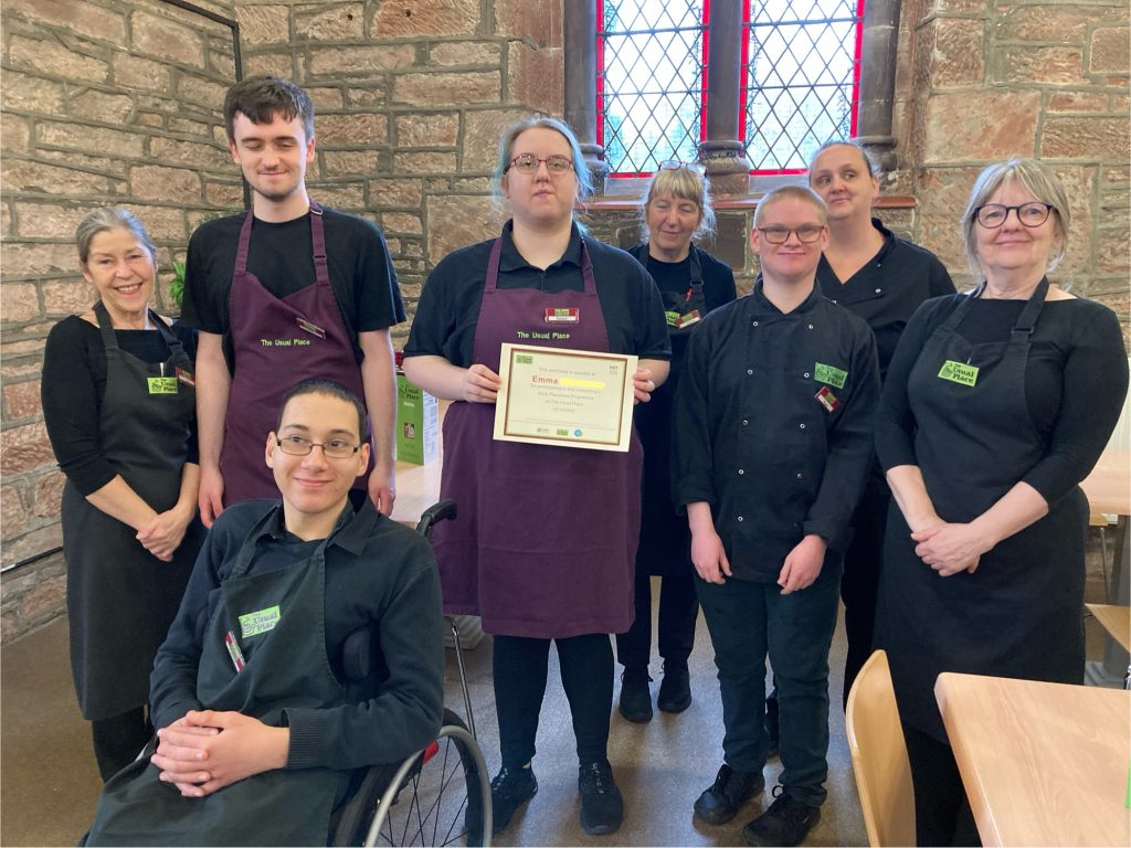 A group of people stand around a young woman in an apron holding a certificate. Everyone is smiling at the camera and wearing The Usual Place blacks and aprons