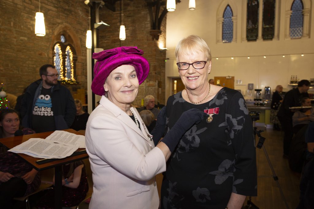 Smartly dressed lady in pink hat stands next to a tall middle-aged lady with short blonde hair and glasses. The former is wearing black gloves and is pinning a British empire Medal with red ribbon to the blonde lady's black dress