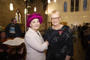 Smartly dressed lady in pink hat stands next to a tall middle-aged lady with short blonde hair and glasses. The former is wearing black gloves and is pinning a British empire Medal with red ribbon to the blonde lady's black dress