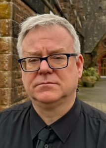 Close up image of a man's face. He is wearing a black shirt and black tie, he has grey hair and glasses. He is standing outside a red sandstone building