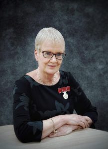 Lady with short blonde hair and glasses sits with arms folded on a table. She is wearing a black dress and on it is pinned a golden British Empire Medal with a red ribbon.
