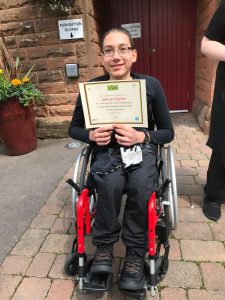 A volunteer in a wheelchair in the centre of the photo wearing The Usual Place uniform. The entrance to The Usual Place is behind him. He is holding a certificate, looking into the camera and smiling.