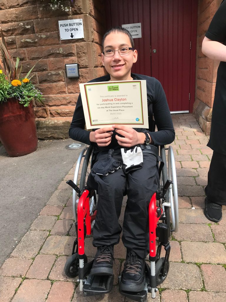 A volunteer in a wheelchair in the centre of the photo wearing The Usual Place uniform. The entrance to The Usual Place is behind him. He is holding a certificate, looking into the camera and smiling.