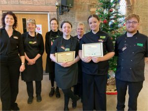 2 young volunteers centre of the photo wearing The Usual Place uniform. Behind them are 4 mentors and 1 volunteer, wearing The Usual Place uniform. The volunteers centre of the photo are holding a certificate each. All of them are smiling into the camera.