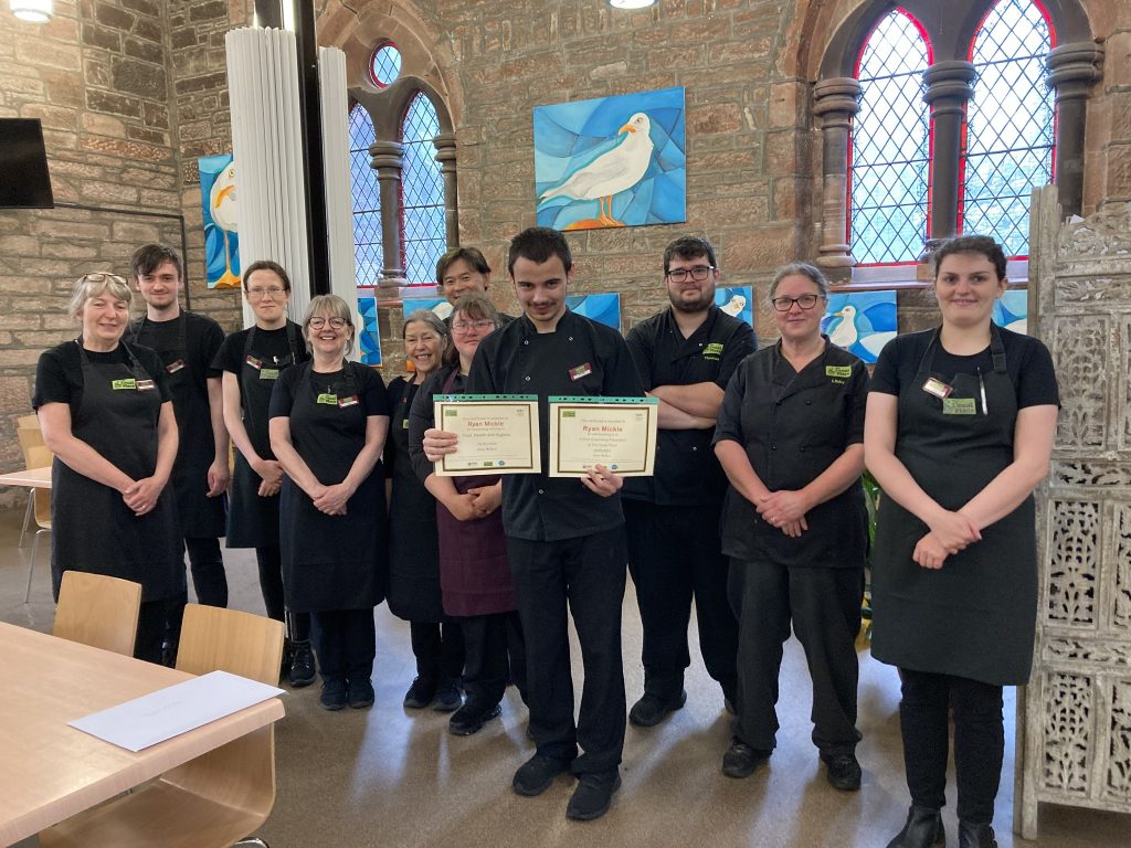 One of the volunteers standing in the centre of the photo wearing The Usual Place uniform. He is holding 2 certificates. Behind him are 3 other volunteers and 7 mentors and staff, also wearing The Usual Place uniform. They are all looking into the camera and smiling.