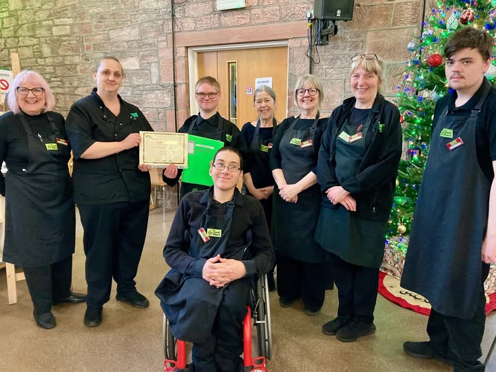 A young person volunteer with glasses standing in the centre of the photo. There is 1 volunteer alongside 1 kitchen mentor on his right, and 3 front of house mentors and 1 volunteer on his left, as well as 1 volunteer in a wheelchair in front of him. All of them are wearing The Usual Place uniform. The kitchen mentor on the left is holding a certificate to give to the one with glasses behind the one in the wheelchair. Everyone is looking into the camera and smiling.