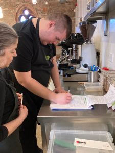 A volunteer is looking down at a clipboard. He is holding a pen and is ticking off items on the clipboard. A mentor stands in front of him. She is watching as the volunteer signs the checklist.