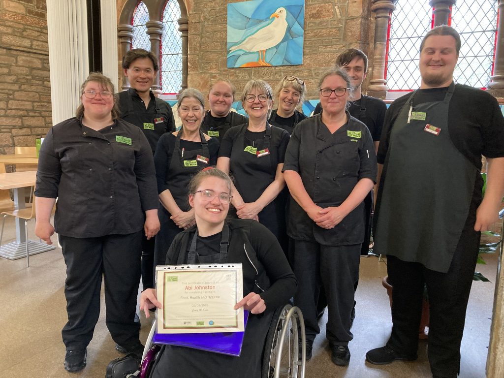 A volunteer in a wheelchair is sat centre of the photo wearing The Usual Place uniform holding a certificate for food safety and hygiene. Behind her stands 5 mentors and 3 volunteers, also wearing The Usual Place uniform. They are all looking into the camera and smiling.