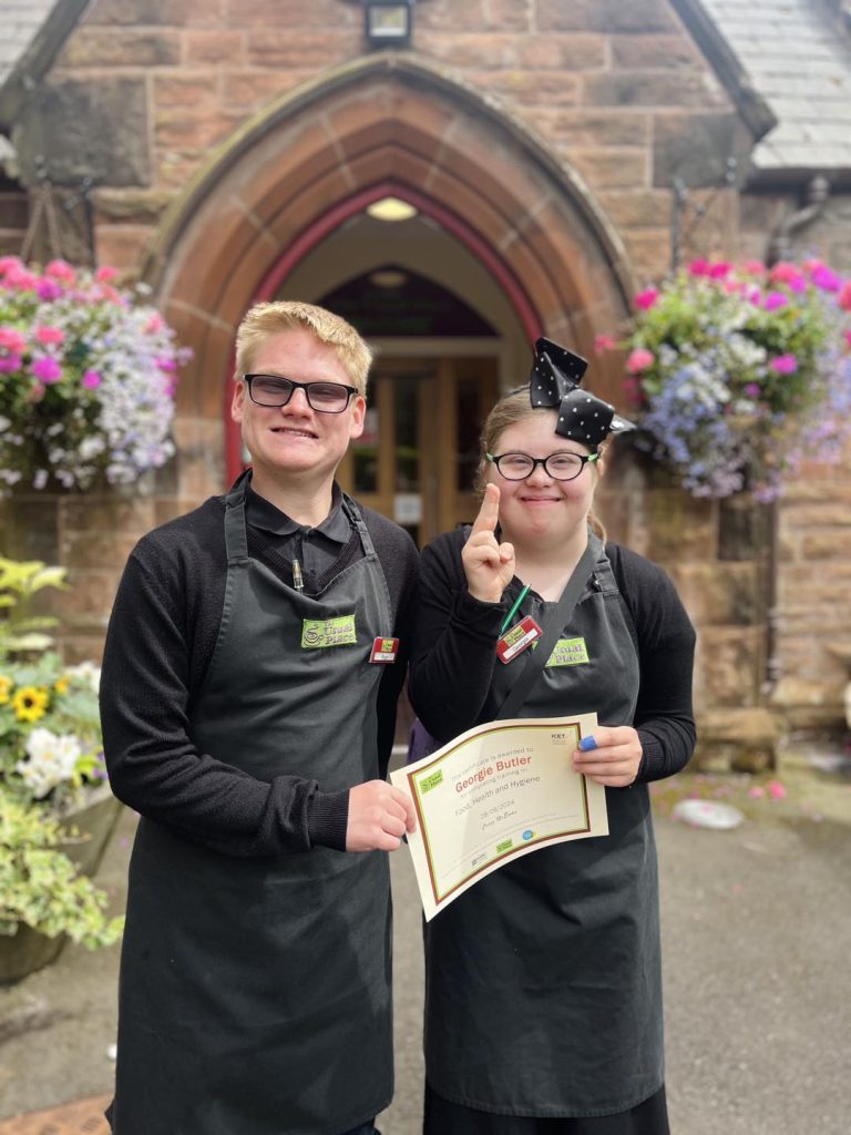 A volunteer with glasses and a bow stood centre-right in the picture wearing The Usual Place uniform. Stood next to her centre-left in the picture is another volunteer presenting a certificate for food, health and hygiene to her. They are both looking into the camera and smiling.