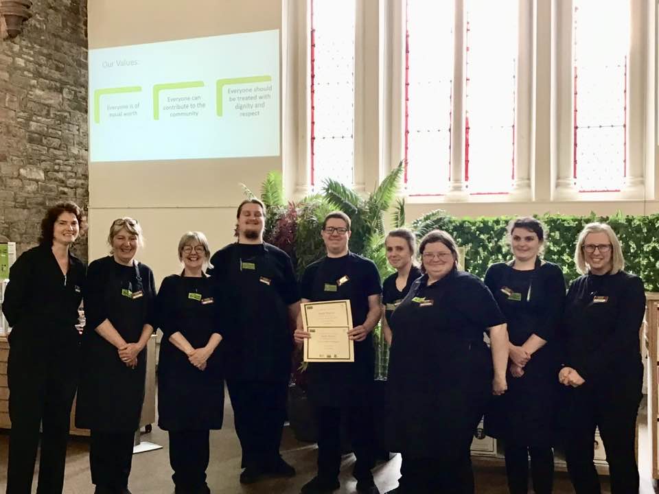 A volunteer with glasses standing in the centre of the photo wearing The Usual Place uniform. He is holding a certificate for food health and hygiene. To the left of him are 3 mentors and a volunteer, and to the right is 1 mentor and 3 volunteers. They are all looking into the camera and smiling.