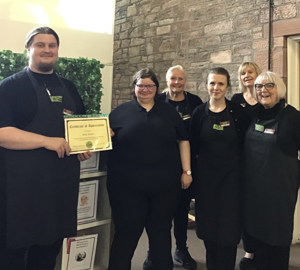 A volunteer with glasses standing in the centre of the photo. There are two more volunteers on her right and her left, and 3 mentors behind them. All of them are wearing The Usual Place uniform. The volunteer on the left is holding a certificate to give to the one with glasses. Everyone is looking into the camera and smiling.