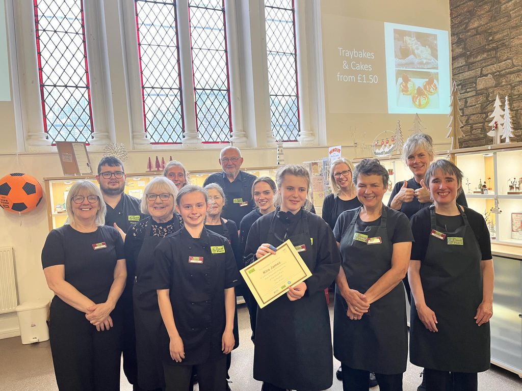 A volunteer with brown hair tied up in a bun standing centre-left of the photo. She is surrounded by staff and other volunteers. One of the other volunteers is holding a certificate for her. They are all looking into the camera and smiling.