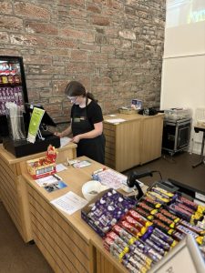 A volunteer with glasses standing centre-right of the picture wearing The Usual Place uniform. She is stood behind the welcome station using the register.